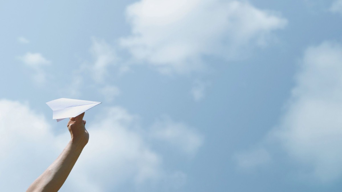 Hand hält einen Papierflieger vor Himmel mit Wolken