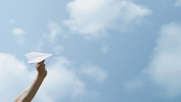 Hand hält einen Papierflieger vor Himmel mit Wolken
